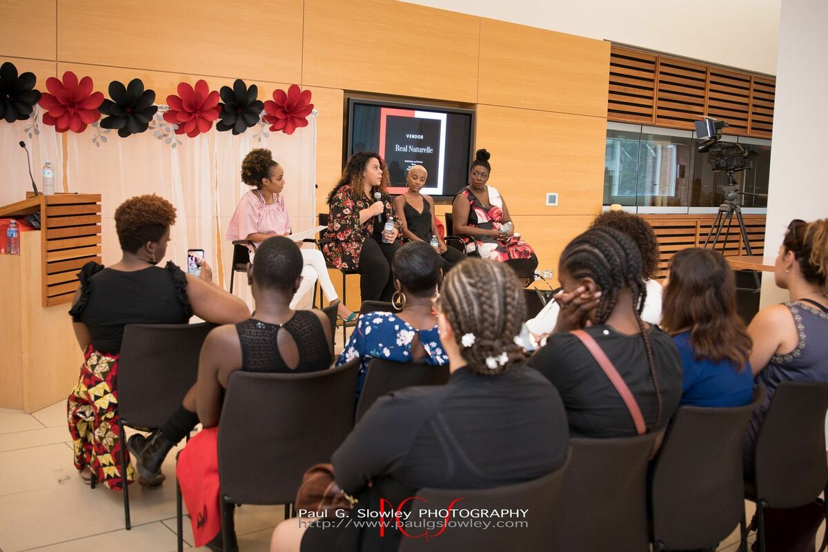 Four Black female panelists sit on a stage speaking to an audience of women
