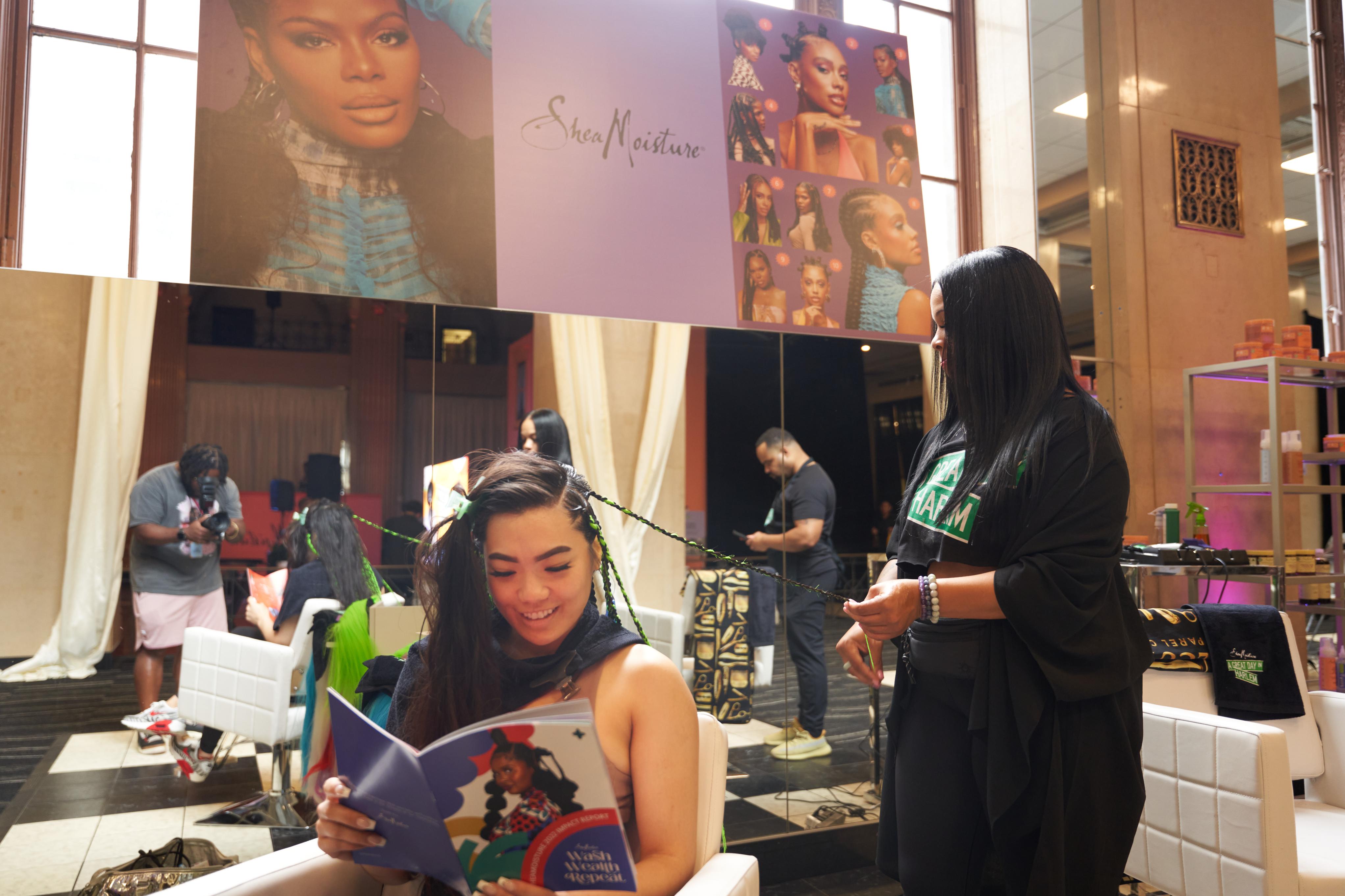 A Black woman at Essence Fest braiding another woman's hair who is reading a book 