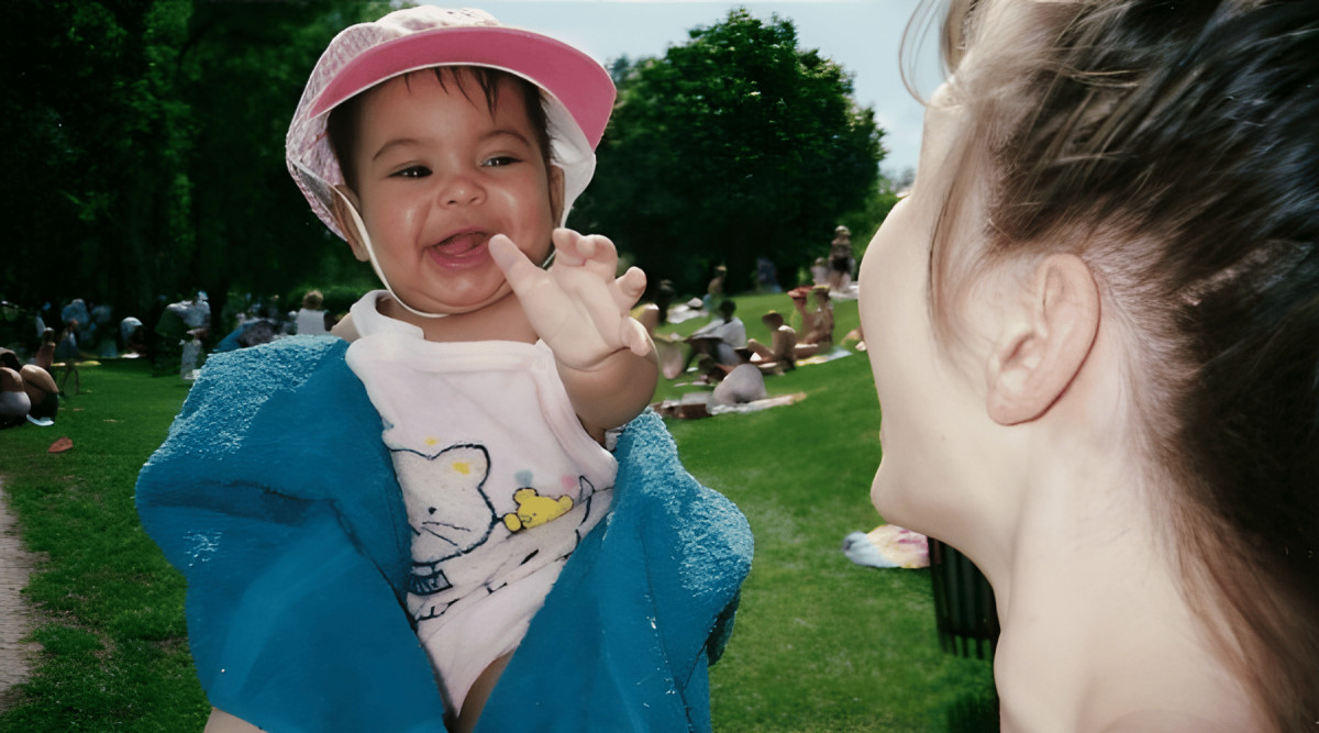 A close-up photograph shows an infant being held outdoors in a sunny park. The baby faces the camera, smiling with one hand raised near their mouth, and wears a pink sun hat, a light-colored shirt with a cartoon graphic, and a bright blue towel or blanket around the shoulders. At the right, part of an adult’s face and tied-back brown hair is visible in profile, facing the baby. Behind them are green grass, leafy trees, and several people sitting or walking in the park.
