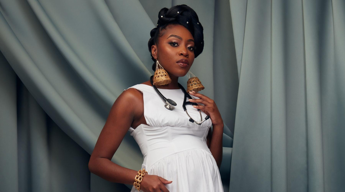 A portrait of a Black woman standing against layered gray-blue draped fabric. She wears a white sleeveless dress with a gathered waist and rests one hand beneath her abdomen while the other touches a black stethoscope hanging around her neck. Her hair is styled in an updo decorated with small metallic accents, and she wears large woven tan earrings and a chunky gold bracelet. She faces the camera with a calm, direct expression.